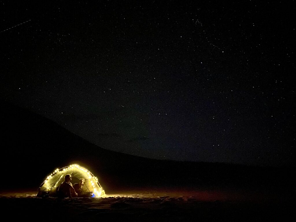 Two people sitting in a tent lit up by lights under the night sky in the Great Sand Dunes National Park 